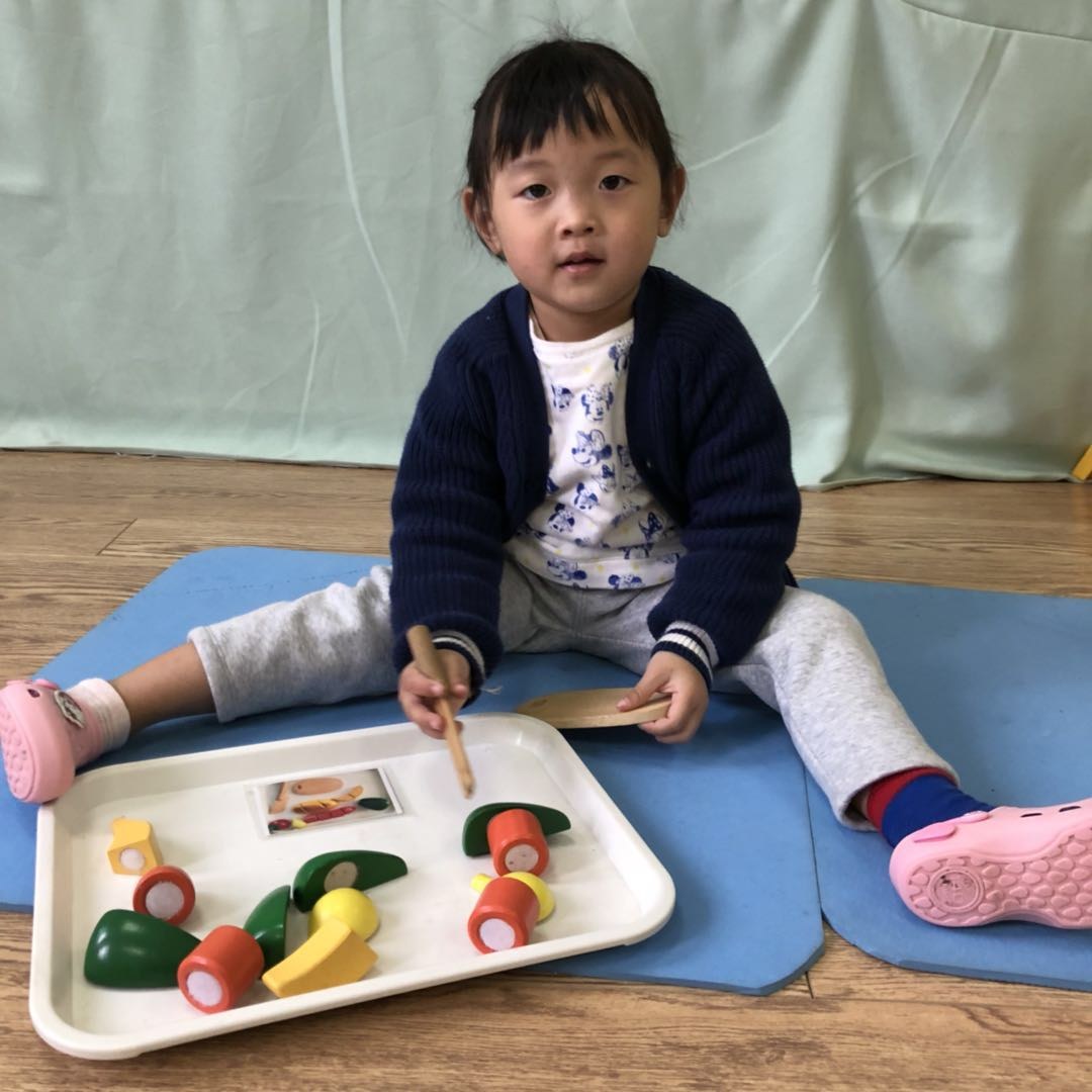 一名幼儿坐在室内软垫上，面前托盘内摆有木制蔬果玩具，手持木棒和木片看向镜头