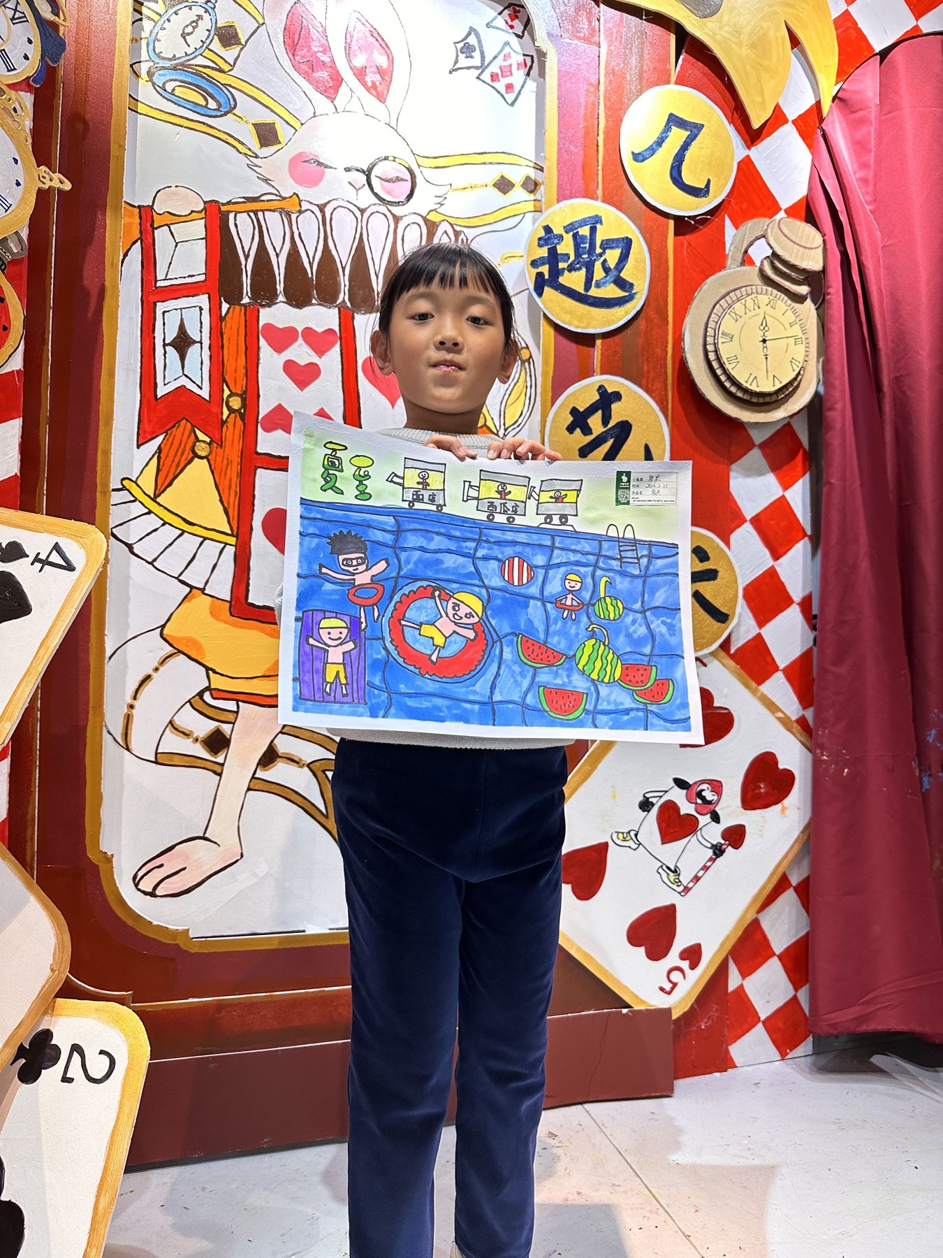 一名女孩站在大型纸艺布景前，双手展示一幅以泳池和西瓜为主题的彩色画作