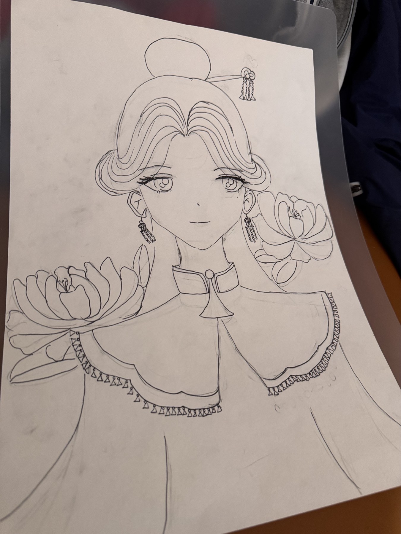 纸张上绘有一幅古装女性半身线稿，人物两侧配有花朵，纸张放在桌面垫板上