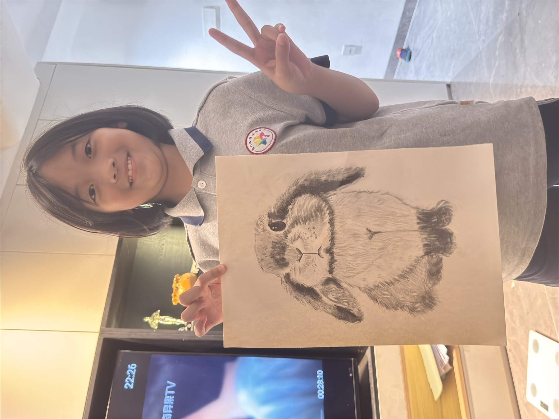 一名女孩在室内举着一幅兔子素描并做胜利手势，身穿带徽标的灰色上衣