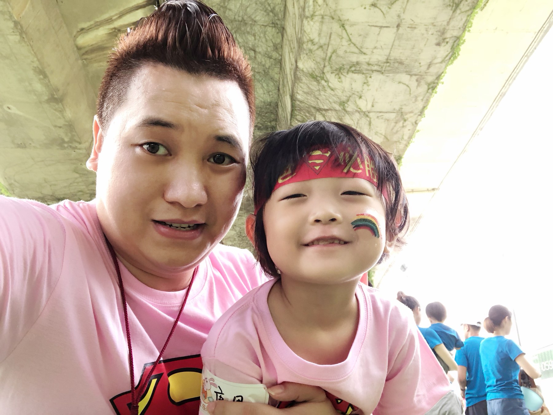 成年男性与幼年女孩近距离自拍合影 女孩佩戴红色头带且脸上有彩绘 背景有多人活动