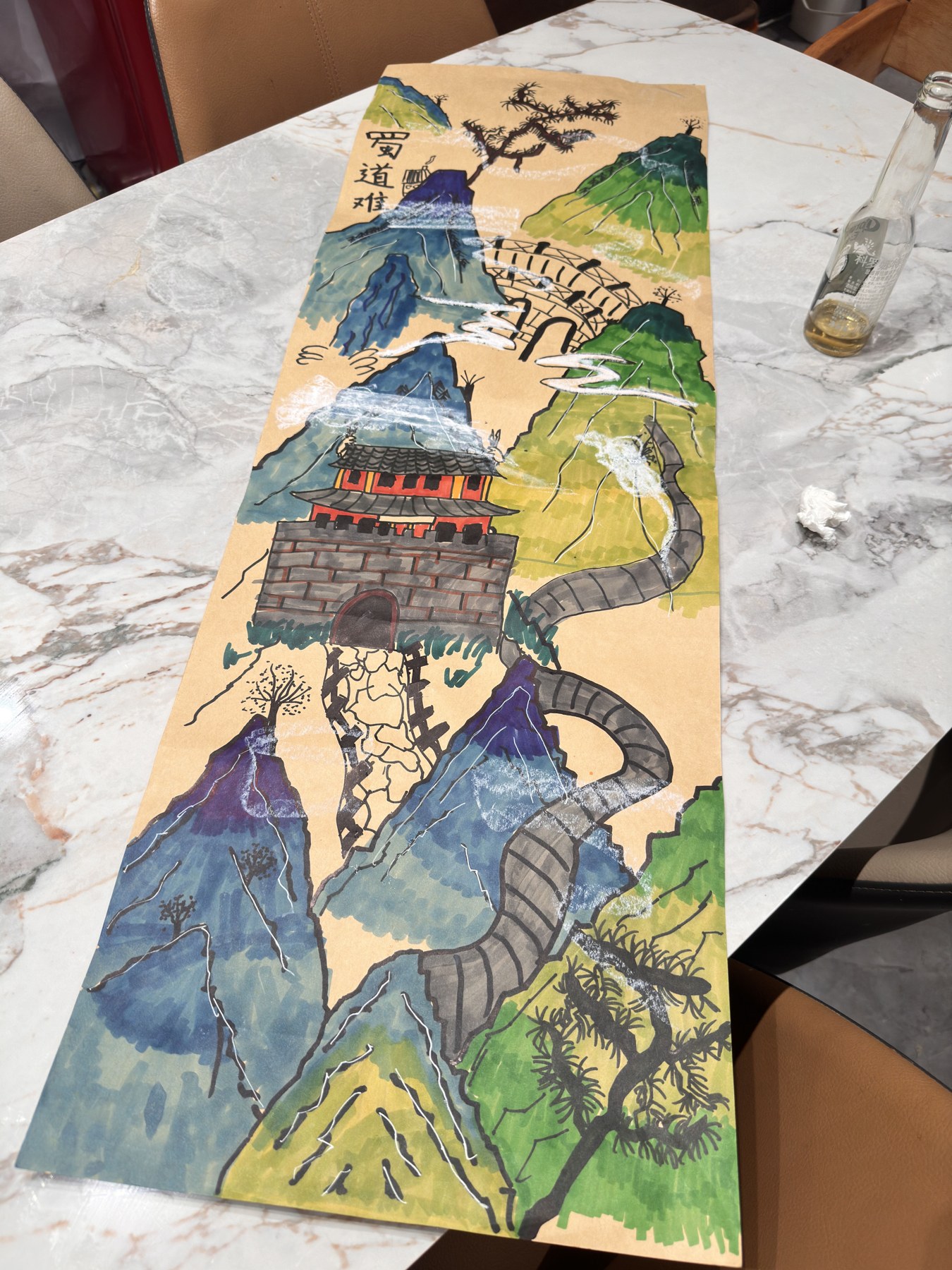 一幅铺在餐桌上的长卷彩色山水画描绘山峰城墙桥梁树木与建筑