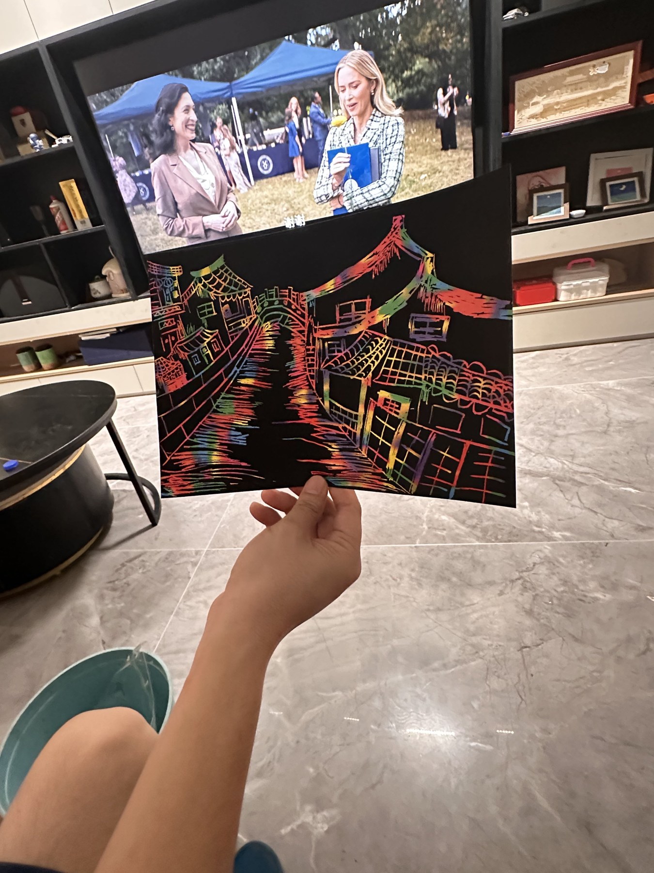一只手在客厅中举着黑底彩色刮画作品，画面描绘临水房屋与桥