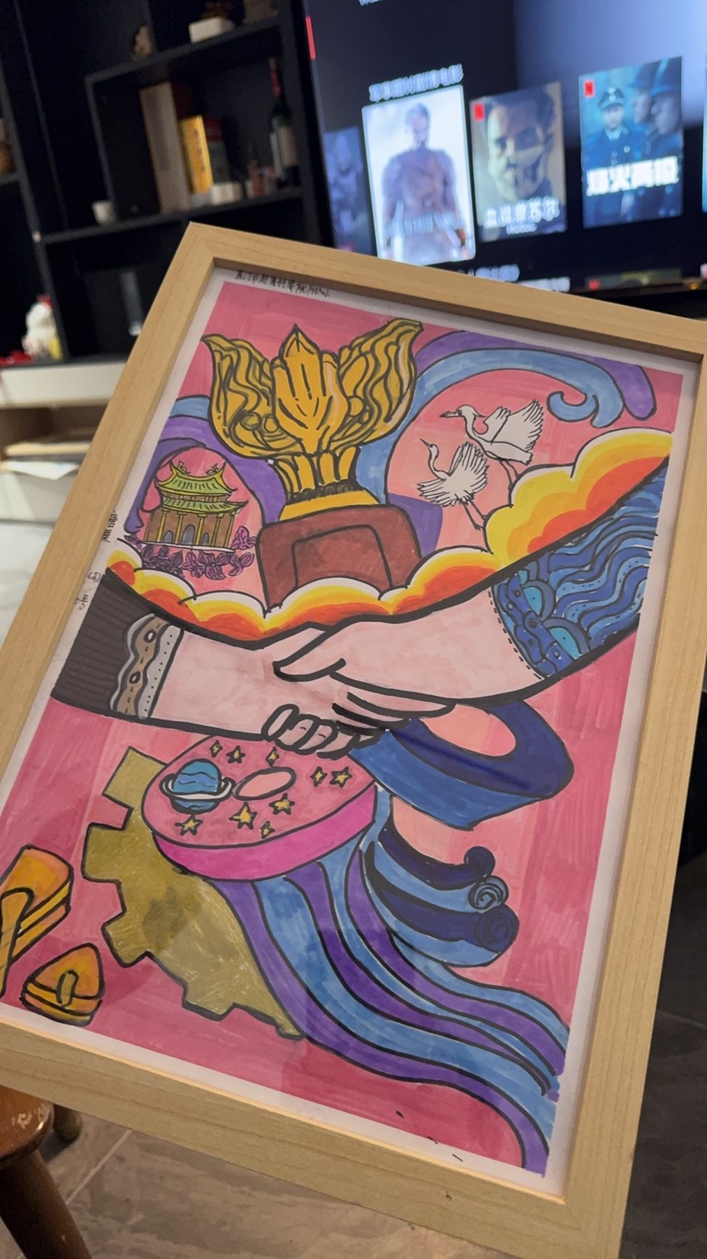 一幅装框彩色画作置于居家空间，画面包含握手、金色花形建筑、飞鸟及城市意象