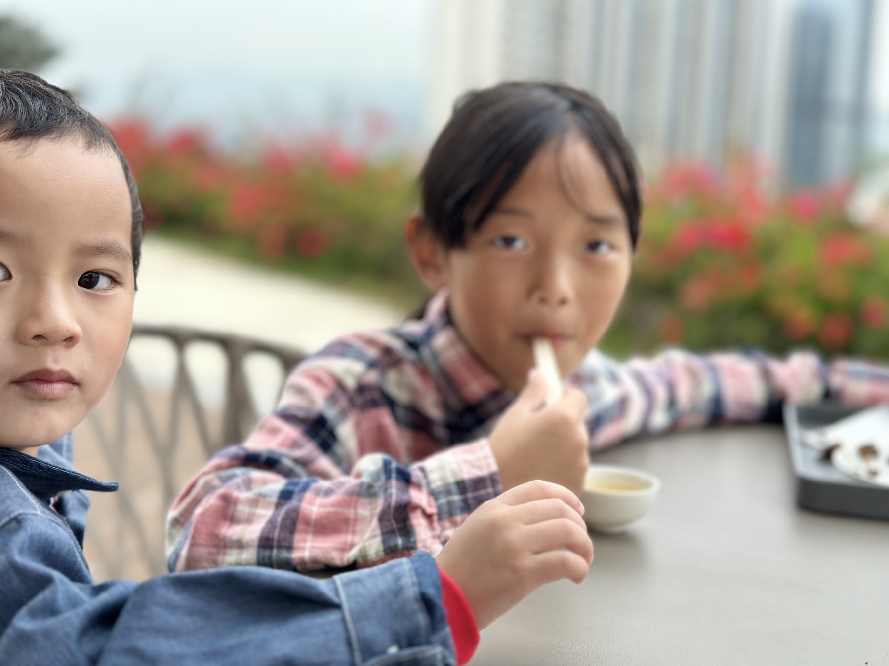 一名幼儿靠近镜头，旁边一名女孩坐在户外桌边进食